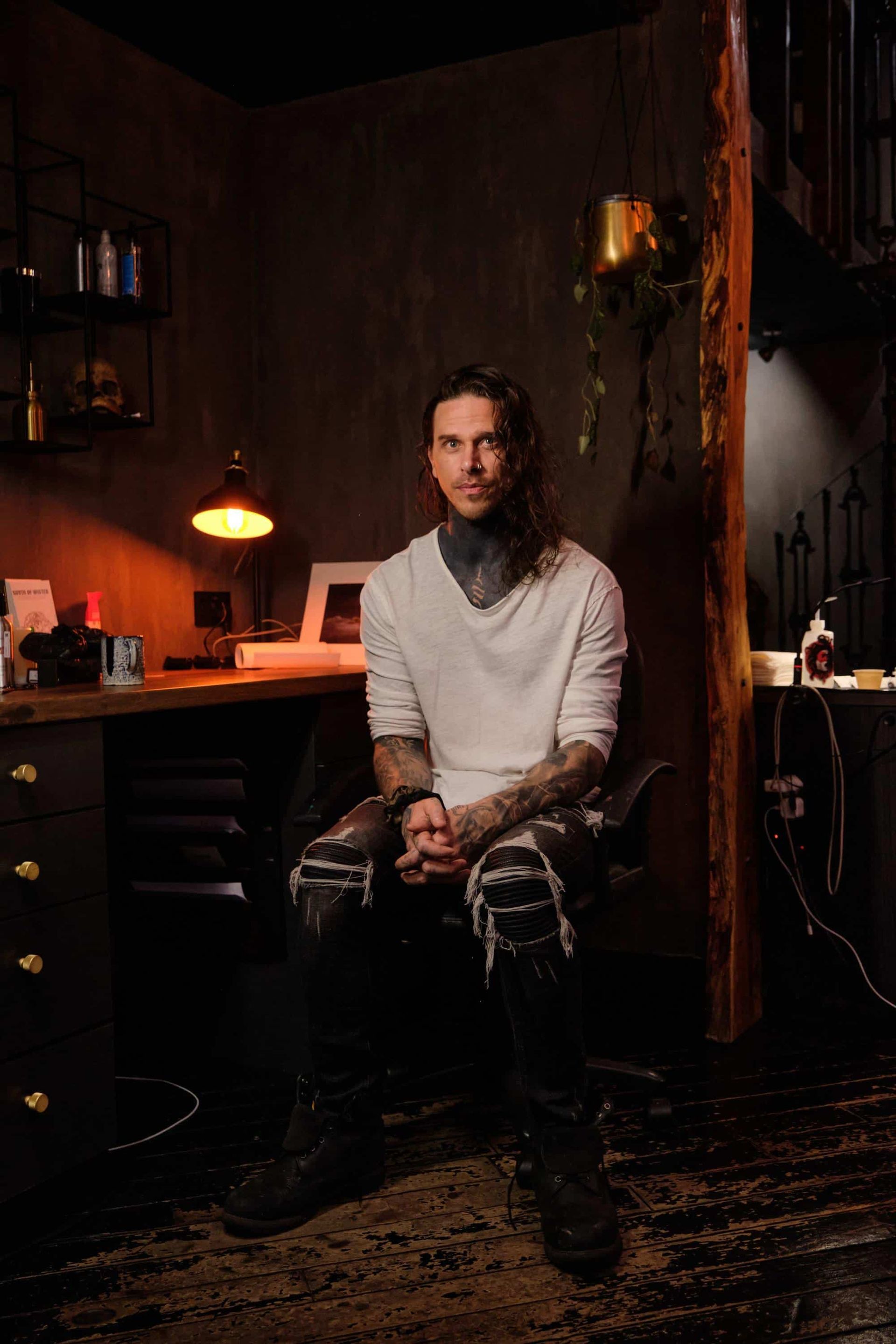 A person with tattoos on their neck and arms sits in a dimly lit, industrial-style studio, wearing a white shirt and jeans.