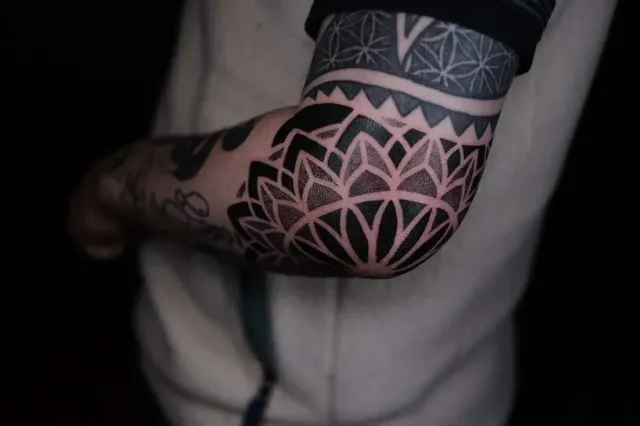 A close-up of a person's elbow featuring a large, black ink mandala tattoo with intricate geometric patterns.