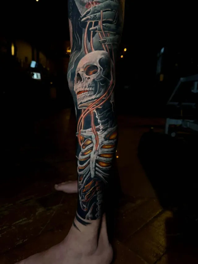 A detailed tattoo of a skull and ribcage on a person's lower leg, featuring glowing orange chains and dark shading.